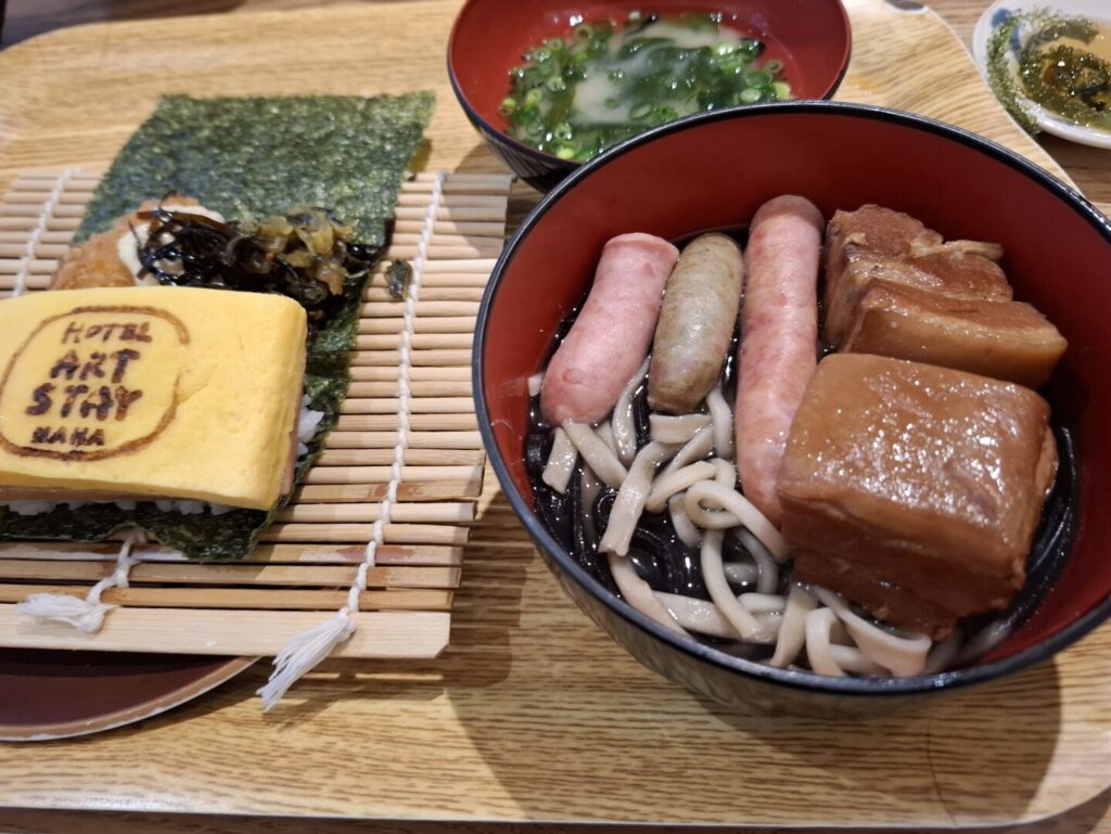 那覇の交際通り近くにある「ホテルアートステイ那覇国際通り」に宿泊した際に撮った朝食の麺料理にソーセージとラフテーを乗せたお椀とポーたまの写真
